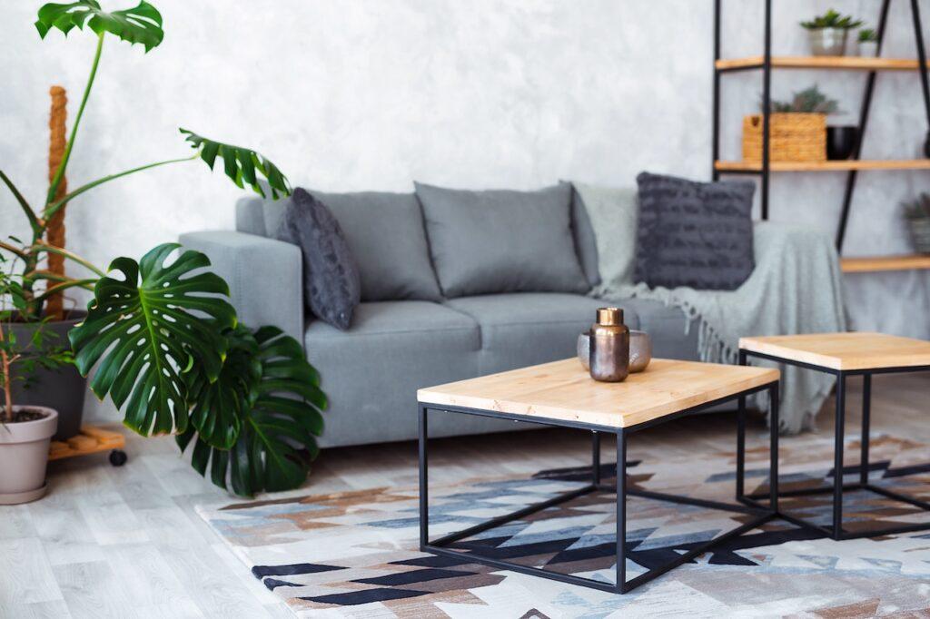 Moderner Couchtisch mit Holzplatte und schwarzem Metallgestell in einem Wohnzimmer mit grauem Sofa, Monstera-Pflanze und Regalen.