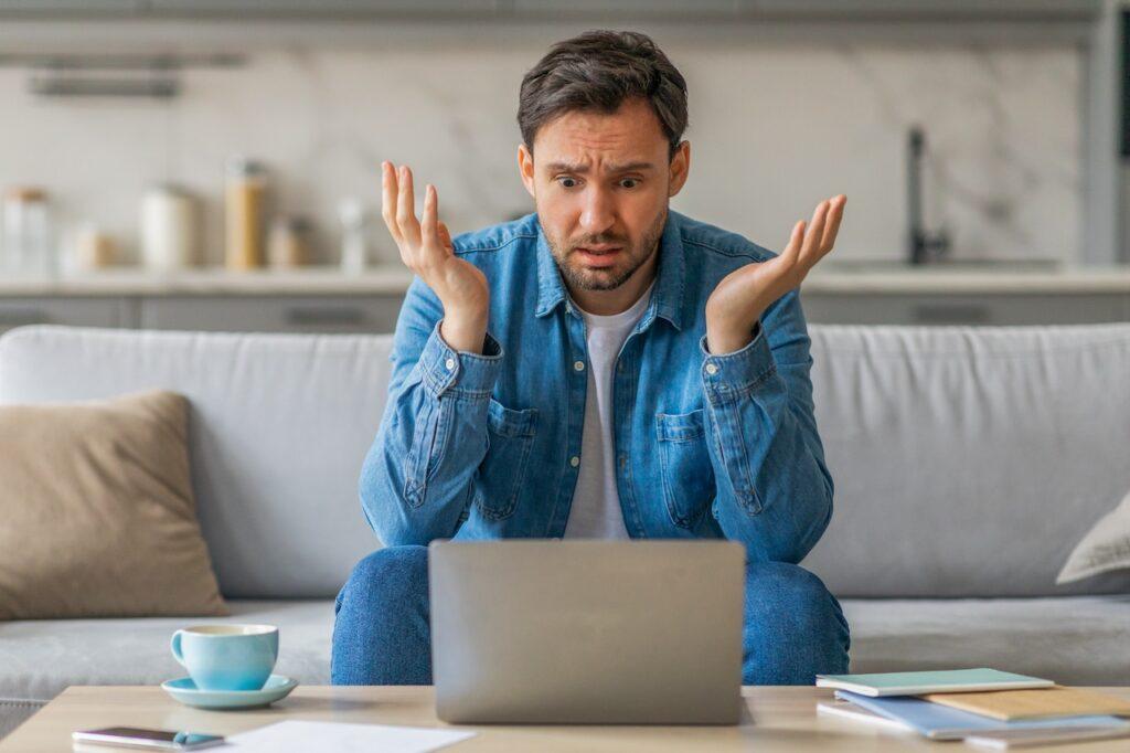 Ein Mann sitzt auf einem Sofa vor einem Couchtisch, schaut verzweifelt auf seinen Laptop, umgeben von einer Kaffeetasse und Dokumenten.