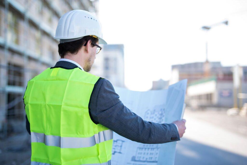 Mann mit Schutzhelm und Warnweste überprüft Baupläne auf einer Baustelle im Anlagenbau